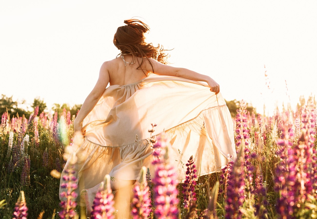 Image featuring a woman strolling through vibrant flower fields in a flowing white summer dress, viewed from behind. Evokes a sense of freedom, purity, and authenticity—mirroring the essence of our natural Champagne.